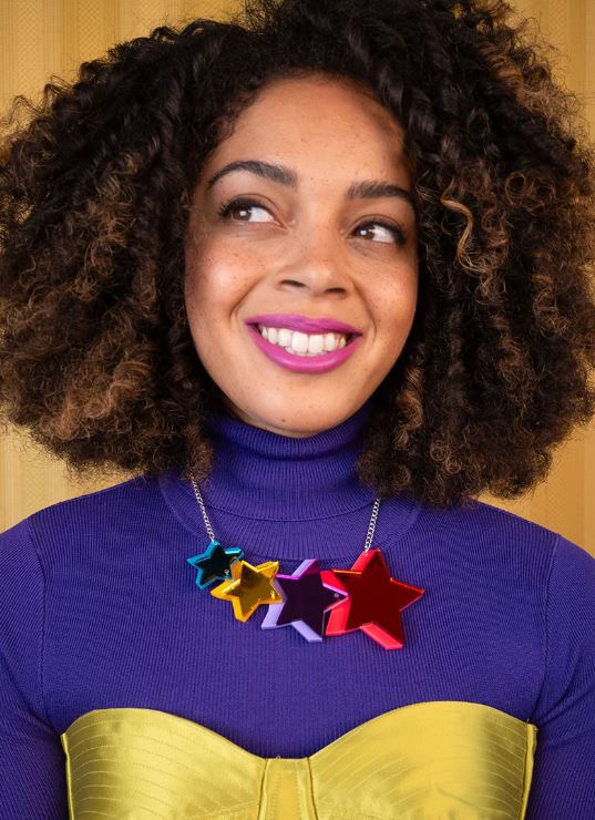 A woman with curly hair smiles while looking to the right, dressed in a purple turtleneck and a striking bright yellow strapless top. She is adorned with a large, colorful Blue Dog Posters Pop Stars Necklace, and her lips are bright pink. The background features a yellow shade with a subtle pattern.