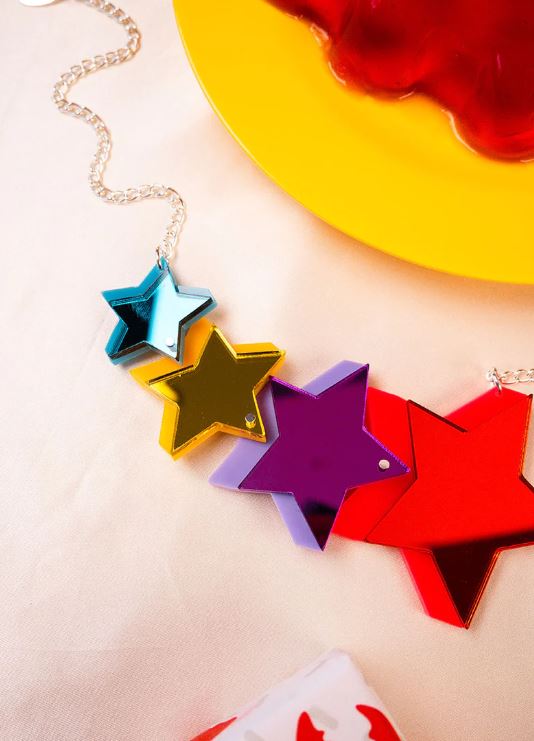 A Blue Dog Posters Pop Stars Necklace featuring colorful star-shaped acrylic charms in blue, yellow, purple, and red lies on a cream-colored surface. Part of a yellow plate with red contents is visible at the edge of the image. This statement necklace adds a playful touch to any outfit.