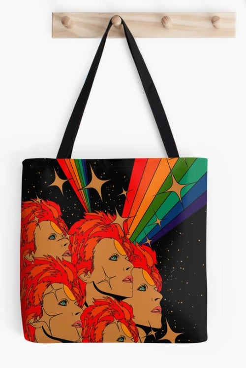 A Starman Tote Bag hanging on a wooden peg rack. Part of our Giftware Collection by Blue Dog Posters, the Audrey Herbertson-designed bag features a colorful design depicting multiple vibrant orange-haired figures gazing upwards. The black background is adorned with stars and rainbow-colored beams radiating from the figures' eyes.