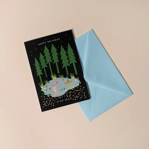 The Stay Wild Swimming Birthday Card by Blue Dog Posters features a pond, trees, and a swimmer, with "Happy Birthday" at the top and "Stay Wild" at the bottom, and comes with a light blue envelope.