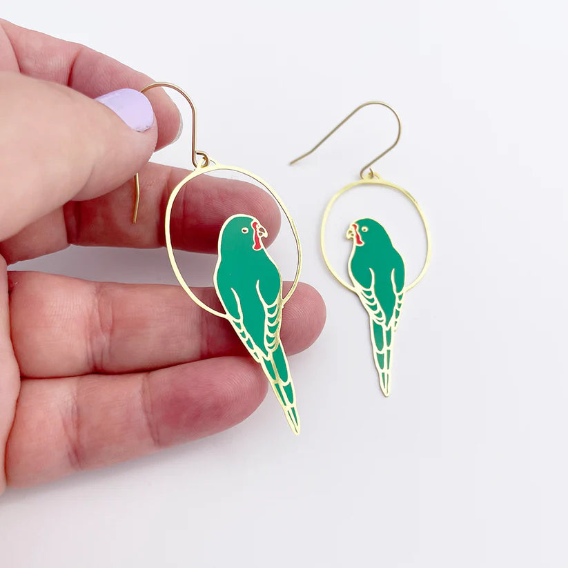 Close-up view of a hand holding a pair of Blue Dog Posters' Parrot in Green Denz Earrings featuring green parrot designs. The parrots hang inside gold hoops and have red beak accents. The person has light purple nail polish on their thumb, showcasing these lightweight Australiana accessories against a plain white background.