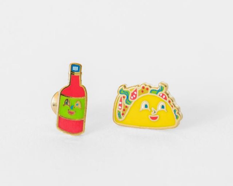 Two vibrant enamel earrings from Blue Dog Posters' Taco & Hot Sauce Earrings collection are set against a white background: one featuring a smiling hot sauce bottle and the other showcasing a cheerful, topped taco. Both earrings are equipped with hypoallergenic posts for comfortable wear.