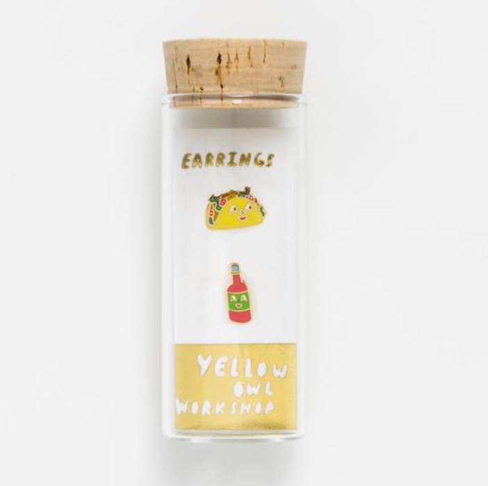 A small clear glass bottle with a cork stopper contains two cloisonné studs: one designed as a taco and the other as a hot sauce bottle, with text below reading "Taco & Hot Sauce Earrings by Blue Dog Posters.