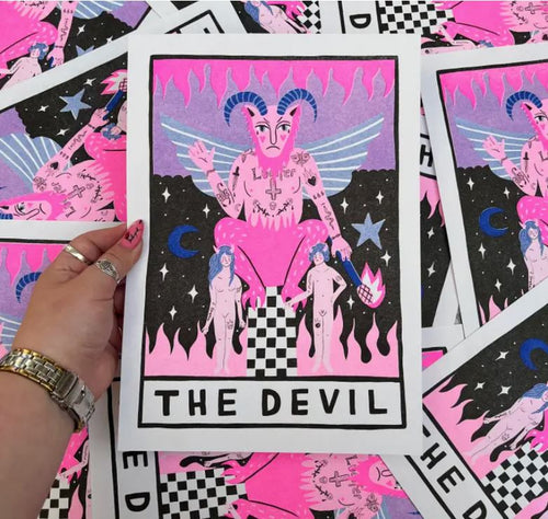 A hand holds the Devil Tarot A4 Risograph by Amy Hastings, featuring vibrant pink and black illustrations. The print, produced by Blue Dog Posters, depicts a devil figure with wings and horns, accompanied by two human figures, flames, stars, and a crescent moon. Multiple copies of this high-quality A4 risograph print are scattered below.