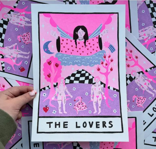 A hand holding a card titled "The Lovers." The card illustration features two nude figures standing below a cloud with an angelic figure above them. Surrounding them are trees, a snake, and a checkered pattern at the top. This high-quality A4 risograph print from Blue Dog Posters, created by Amy Hastings, uses bright pink and purple hues on 170gsm recycled coffee cup paper.