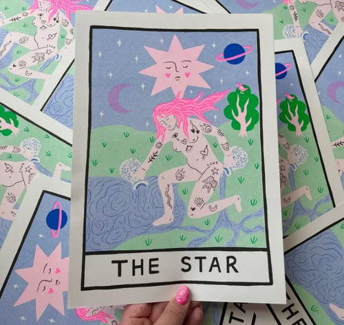 A hand holding a high-quality Star Tarot A4 Risograph print labeled "THE STAR" from Blue Dog Posters. The card, designed by Amy Hastings, features a naked figure with pink hair pouring water from two jugs, adorned with various tattoos. The background includes a pink star with a face, a purple planet, green hills, and a crescent moon. Other identical tarot cards are scattered beneath the primary one. Printed on 170gsm recycled coffee cup paper.