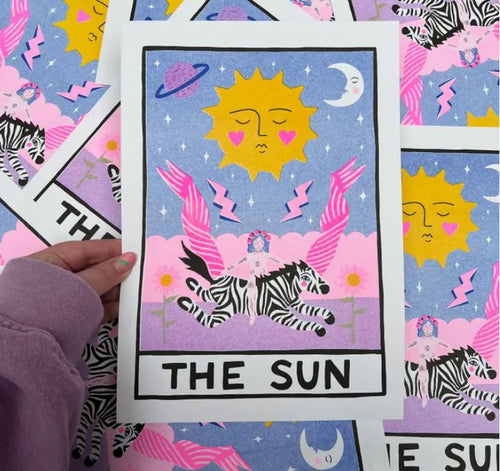 A hand in a purple sleeve holds a colorful tarot card featuring "The Sun." The card, an Amy Hastings Sun Tarot A4 Risograph print by Blue Dog Posters, depicts a sun with a face, pink hearts, a purple planet, a crescent moon, and a black-and-white striped animal with pink wings against a vibrant pink and blue background.