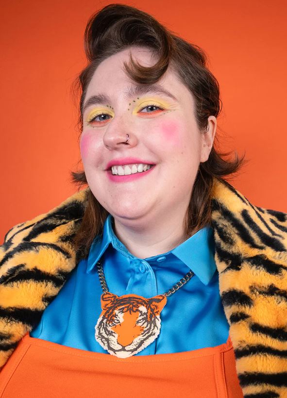 A person with short hair smiles at the camera, adorned with bold yellow eyeshadow, pink blush, and a nose ring. They are dressed in a blue shirt and an orange apron topped with a tiger-print faux fur coat. Completing the ensemble is a Tiger Necklace by Blue Dog Posters made of recycled acrylic, which perfectly complements the vibrant orange background.