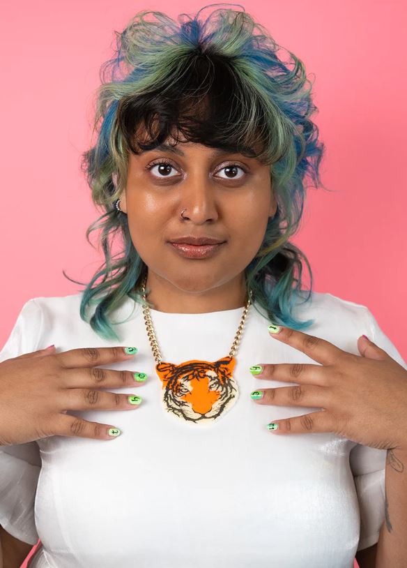 A person with multicolored hair and bangs poses in front of a pink background. They are wearing a white shirt and a Blue Dog Posters Tiger Necklace made of recycled acrylic. Their nails are painted green with black-and-white designs as they gently touch the necklace with both hands.