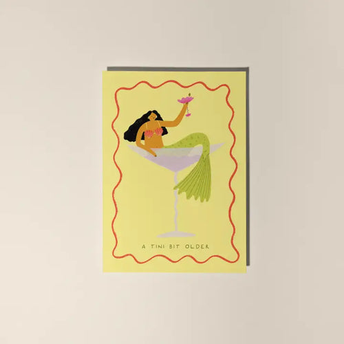 The "A Tini Bit Older Mermaid Birthday Card" by Blue Dog Posters features a woman dressed as a mermaid lounging in a cocktail glass, holding a drink, with a wavy red border and "A TINI BIT OLDER" text at the bottom.