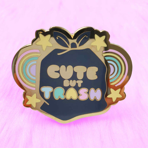 The Trash Bag Pin Pinstachiio, crafted by Blue Dog Posters, is a vibrant enamel pin featuring a black trash bag shape in the center with the phrase "CUTE BUT TRASH" inscribed on it. Encircling the text are pastel-colored rainbows and yellow stars set against a fluffy pink background. The pin is gold plated and includes a back-stamp of authenticity.