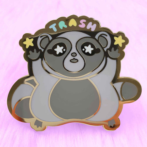The Trash Panda Pin Pinstaciio from Blue Dog Posters is an enamel pin featuring a stylized raccoon with "TRASH" written above its head. The raccoon is raising its arms, with stars in both hands, and the design is accentuated by a shiny gold outline. The pin comes set against a soft, pink, fluffy background and includes a back-stamp of authenticity from the designer Pinstachiio.
