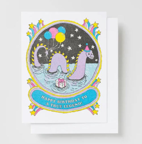 The "Happy Bday True Legend Risograph Card" by Blue Dog Posters showcases two sea serpent characters celebrating a birthday. One serpent is adorned with a birthday hat and clutching balloons, while the other swims nearby holding a gift. The card is printed with veggie-based inks on recycled paper, and features star and wave decorations along with the message, "HAPPY BIRTHDAY TO A TRUE LEGEND!