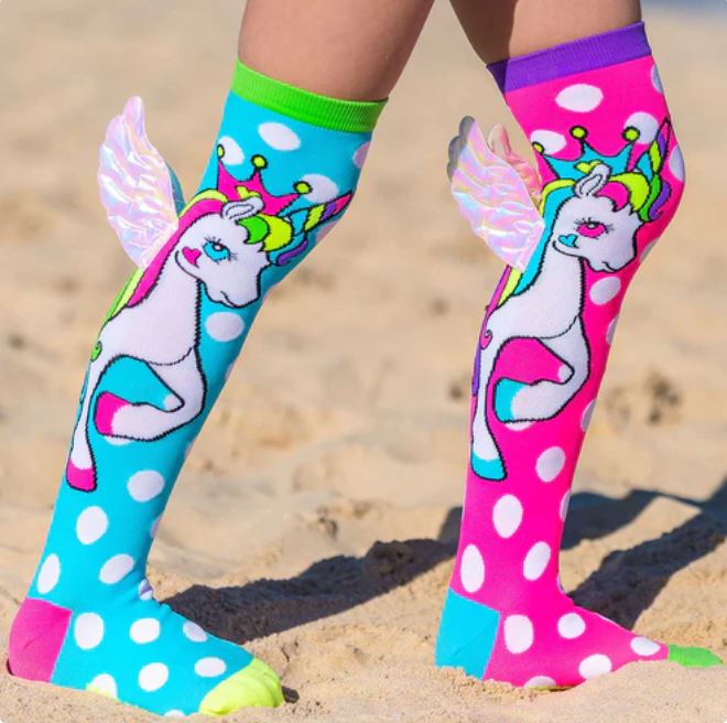 A child stands on a sandy beach, proudly wearing colorful Madmia Flying Unicorn Socks from Blue Dog Posters. These vibrant knee-high socks are adorned with wings and feature polka dots in a playful mix of blue, pink, purple, and green colors. Each unicorn print on the socks sports rainbow-colored manes and tails, creating a delightful mix-and-match look.