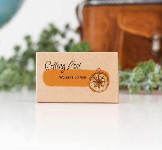 A compact box labeled "Getting Lost Cards - Walkers Edition" by Blue Dog Posters rests on a white surface. The box showcases an illustration of a compass against a blurred backdrop of green foliage and a brown bag, promising a unique walking adventure each time for explorers on foot.