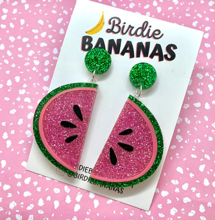 Displayed on a card from "Blue Dog Posters," a pair of summery style earrings designed like watermelon slices with pink and green glitter are featured. Handmade in Australia, the "Watermelon Acrylic Earrings Blue" have green glittery round studs connected to the pink and green watermelon slices. The background is pink with white speckles.