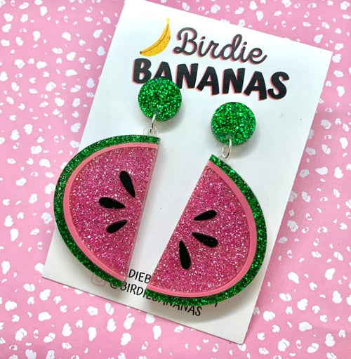 Displayed on a card from "Blue Dog Posters," a pair of summery style earrings designed like watermelon slices with pink and green glitter are featured. Handmade in Australia, the "Watermelon Acrylic Earrings Blue" have green glittery round studs connected to the pink and green watermelon slices. The background is pink with white speckles.