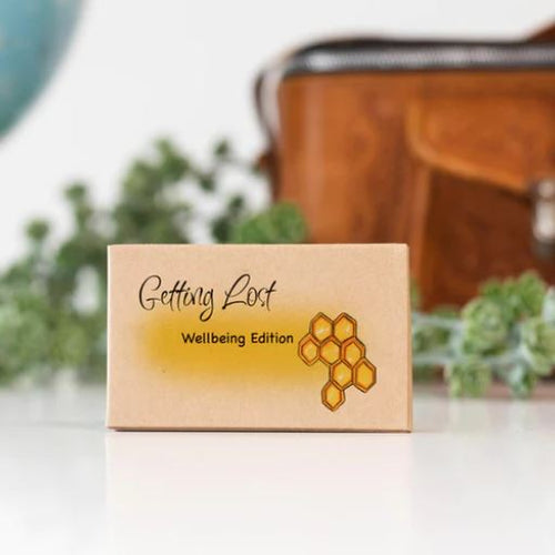 A cardboard box labeled "Getting Lost Cards - Well Being Edition" from Blue Dog Posters is displayed, promising a mindful wellbeing boost. The box features a honeycomb design, with a blurred plant and brown leather bag in the background.