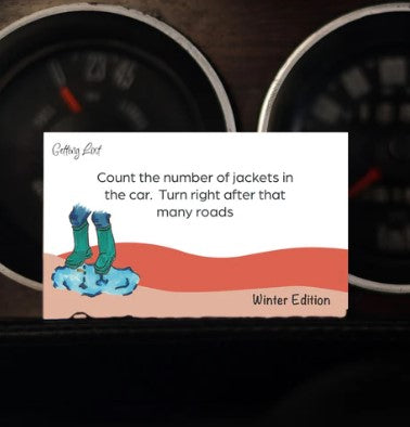 A card on a car dashboard displays instructions: "Count the number of jackets in the car. Turn right after that many roads." This Getting Lost Card, labeled "Winter Edition" by Blue Dog Posters, features an illustration of boots with water pooled around them—a useful tool for adventurers exploring places this season.