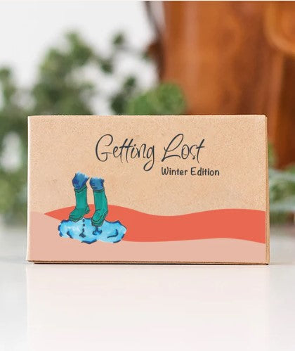 A rectangular beige box features the Blue Dog Posters' "Getting Lost Cards - Winter Edition" written in a stylized font. Below the text, there's an illustration of a snowman lying down on a stylized red snow drift. Designed for winter adventurers, the background includes a blurred brown object and some green foliage.