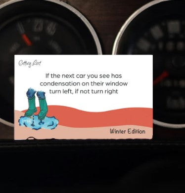 A "Getting Lost Cards - Winter Edition" from Blue Dog Posters sits on a car dashboard. It reads, "If the next car you see has condensation on their window turn left, if not turn right." An illustration of feet in green socks standing in snow graces the bottom, perfect for adventurers exploring places this winter.