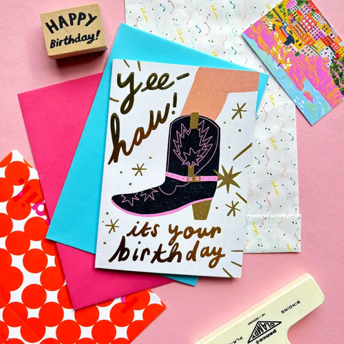 The Yee Haw It's Your Birthday Greeting Card by Blue Dog Posters features a cowboy boot and “Yee-haw! it’s your birthday” text, shown on pink and blue envelopes with festive wrapping paper and a "Happy Birthday!" sign.