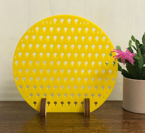 The Round Earring Stand Yellow from Blue Dog Posters, a yellow, circular decorative piece with small cut-out keyhole shapes, is displayed stylishly on a wooden stand. It rests on a wooden surface next to a potted cactus with a pink flower, highlighting its simple and functional design as an earring stand.