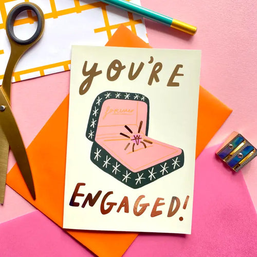 The You're Engaged Ring Greeting Card by Blue Dog Posters features an open ring box with a diamond ring and bold "YOU'RE ENGAGED!" text, surrounded by scissors, a pencil, tape, and colorful envelopes.