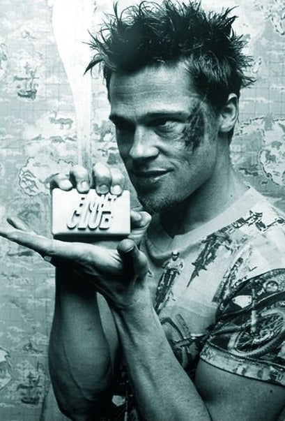 A man with spiky hair and a bruised face holds a bar of soap with "Fight Club" written on it. He is wearing a graphic T-shirt, and the image has a gritty, monochromatic filter. The background is a distressed wall, giving this Brad Pitt Fight Club Poster its definitive Cult Movie Poster charm by IMP.