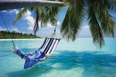 A hammock draped with a colorful blanket swings under a palm tree over crystal-clear turquoise water. The sky is bright blue with a few clouds, and a sandy beach with lush greenery is visible in the background—a perfect scene for your Beach - Morning from our NAT Scenic Collection.