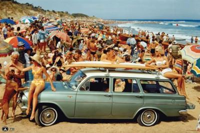 A bustling beach scene features numerous people lounging under vibrant umbrellas. Parked on the sand is a nostalgic Holden - Beach Vibes station wagon by CAR 008, reminiscent of a Motor Sport Poster. Two surfboards rest atop the car, while sunbathers in swimwear gather around and on the vehicle, soaking up the sunshine.