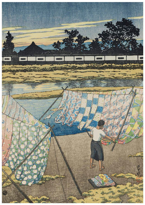 A person hangs a variety of colorful fabrics on a line to dry by a riverbank under a partly cloudy sky. Trees and a white building with a dark tiled roof are visible in the background, reminiscent of an exquisite A3 490 Evening at the Kamo River printed on high-quality 250gsm Satin Paper.