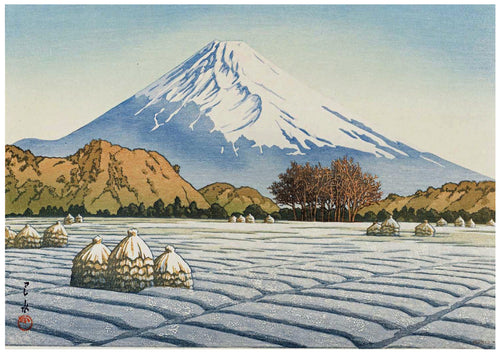 A serene winter landscape features Mount Fuji in the background, its peak covered in snow. In the foreground, snow-covered straw bundles dot a frozen field. Leafless trees stand to the right, with rolling hills separating the mountain from the fields. Perfect as an A3 492 Frosty Morning Japanese Art Poster on high quality 250gsm Satin Paper.