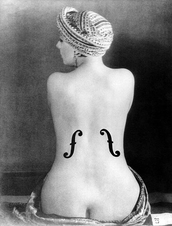A black and white photo of a person sitting bare-backed, facing away from the camera. The person's head is covered with a patterned headwrap, and there are violin f-holes painted symmetrically on their back, resembling the body of a string instrument. This image is titled "Le Violon d Ingres 1924" by IMP 090.