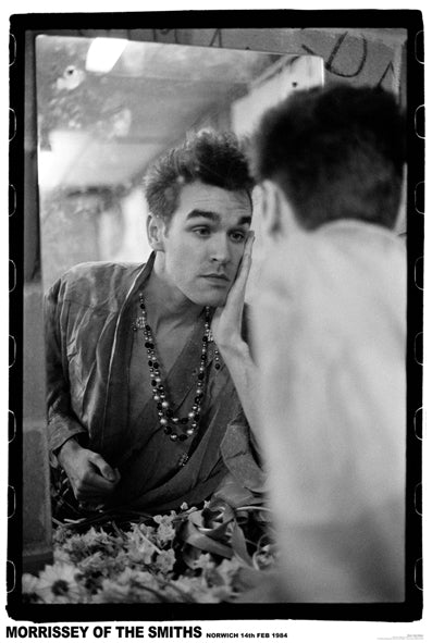 Black-and-white photo of a man with short hair looking at his reflection in a mirror. He rests his face in one hand and wears a beaded necklace over a loose shirt. The bottom caption reads, "Morrissey of The Smiths, Norwich 14th Feb 1984." Ideal for music posters printed on 150gsm gloss paper. Product name: The Smiths Norwich 1984 by MUS 080.
