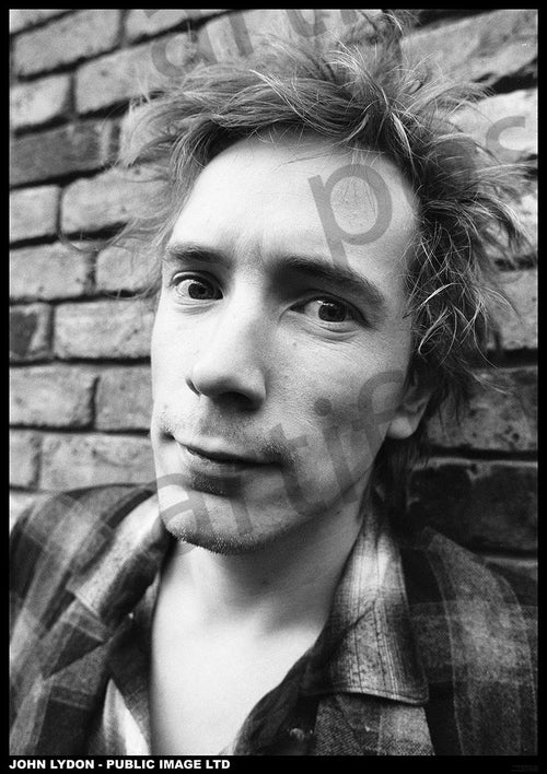 Black and white photo of John Lydon with spiky hair standing against a brick wall. He is wearing a plaid shirt and looking directly at the camera with a slightly tilted head, making it perfect as a rock poster for your music collection. Available from MUS 145, this "John Lydon Public Image LTD" poster is printed on premium 150gsm gloss paper.