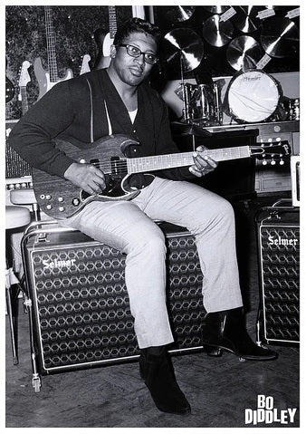 A man sits on a speaker cabinet, engrossed in playing an electric guitar. Behind him, a variety of instruments such as drum kits and additional guitars can be seen. The image, rendered in black and white, is printed on premium gloss paper and features the text "Bo Diddley London" at the bottom right corner—ideal for any rock poster music collection from MUS 136.
