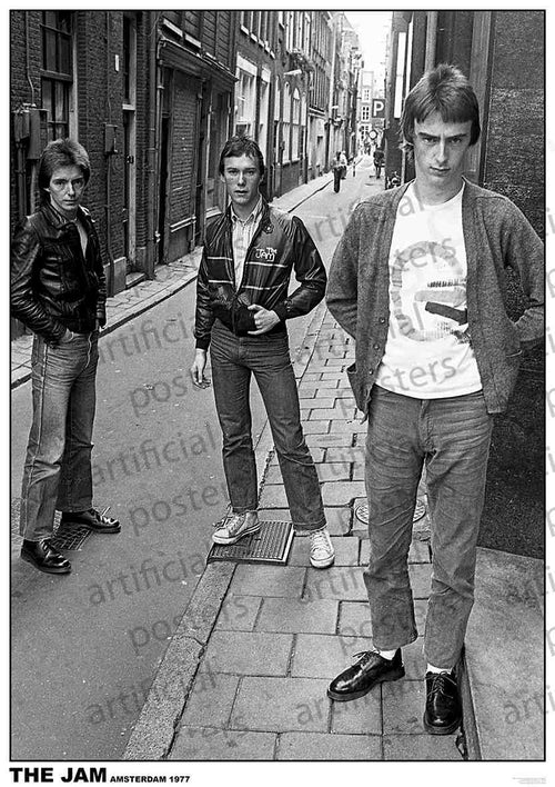 Black and white image of three men from the band The Jam standing in an alleyway in Amsterdam in 1977. They are dressed in casual clothes typical of the era, with the man in front wearing a cardigan and t-shirt, and the men behind in jackets and jeans. Perfect addition to any MUS 224 The Jam rock poster music collection.