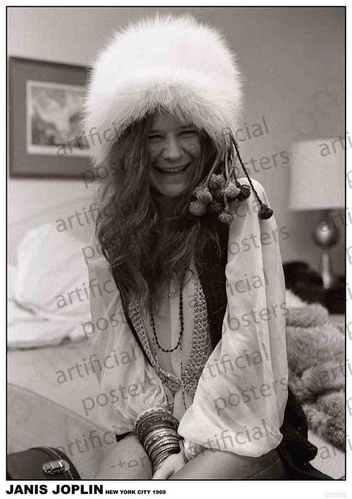A black-and-white photo of a person smiling widely, with long, wavy hair and wearing a fluffy hat, several layered necklaces, and multiple bracelets. Behind them is a bed adorned with a fur blanket and a framed picture on the wall. The text below reads "Janis Joplin New York City 1969." Perfect for any Rock Poster music collection. Product Name: Janis Joplin 1969 by Brand Name: MUS 085.