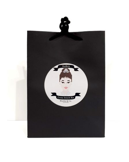 A black gift bag with a circular label featuring a pixelated portrait of a woman with a tiara, pearls, and an updo hairstyle. The label reads "Hustle and Co. Cross Stitch - Audrey" by "Blue Dog Posters." This part of the Cool Gifts Collection also includes an embroidery hoop inside. The bag has black rope handles.