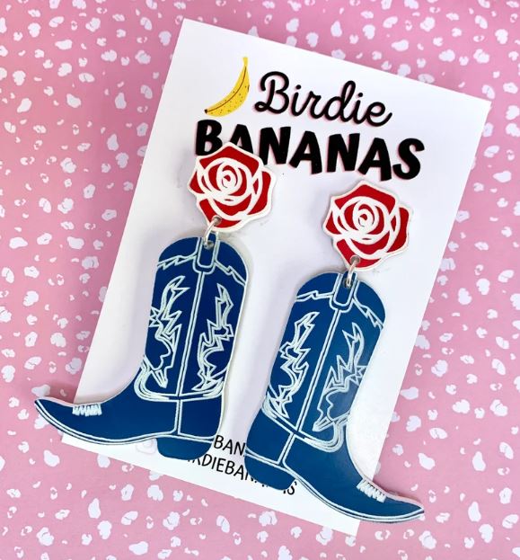 A pair of Cowboy Boots Acrylic Earrings in blue, made by Blue Dog Posters, featuring white detailing and red rose tops is displayed on a card labeled "Birdie Bananas." The background is pink with white speckles, capturing the essence of fun quirky accessories and handmade jewelry.