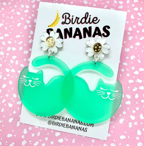 A pair of cheerful green "Curvey Cat" acrylic earrings featuring whimsical white cat illustrations, attached to daisy-shaped earring studs with beige centers. These quirky and fun accessories, crafted by Blue Dog Posters, come displayed on a card labeled "Birdie Bananas," with a pink polka dot background, showcasing unique handmade jewelry.