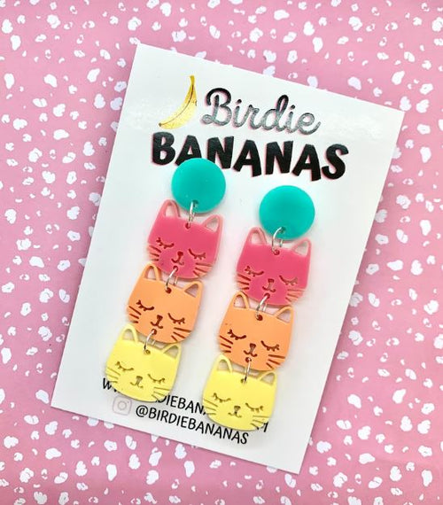 A pair of Kawaii Cat Acrylic Earrings featuring stacked cat heads in pink, orange, and yellow on teal circular studs. Each cat has a different expression. These hand-made jewelry pieces are displayed on a card with the brand name "Blue Dog Posters." The background is a pink surface with white speckles.