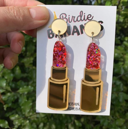 A person holds up a card displaying Lipstick Acrylic Earrings from Blue Dog Posters, designed to look like lipsticks. These fun, quirky accessories feature gold bases and glittery red tops. The card, branded "Blue Dog Posters," is set against a background of green foliage.