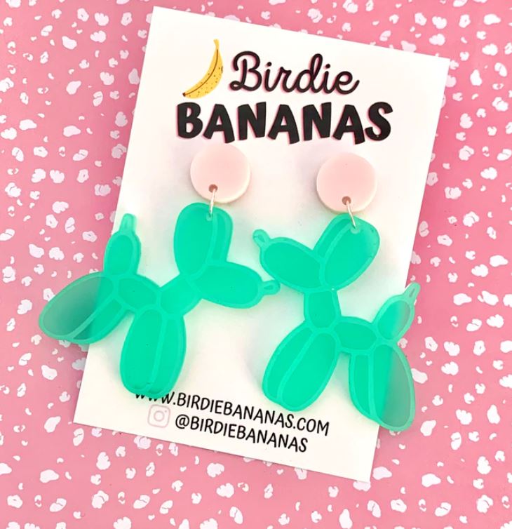 Earrings shaped like green balloon animals are displayed on a card that features "Blue Dog Posters" at the top. The card, set against a pink background with white speckles, showcases these fun and quirky accessories which have pink circular studs. The website and Instagram handles are also provided at the bottom.