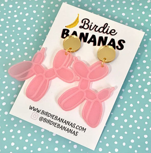 A pair of Balloon Dog Acrylic Earrings in pink, complete with golden stud tops, is displayed on a card featuring the "Blue Dog Posters" branding. This delightful piece of hand-made jewellery rests on a light blue polka-dot background, with contact details for Blue Dog Posters printed at the bottom.