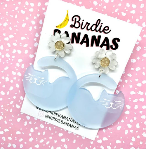 A pair of fun, quirky Curvey Cat Acrylic Earrings Blue from Blue Dog Posters is displayed on a pink speckled background. Each handmade earring features a white floral stud with a yellow center and a dangling light blue cat shape with engraved facial details. The card behind the accessories showcases the text "Birdie BANANAS" with a banana graphic.