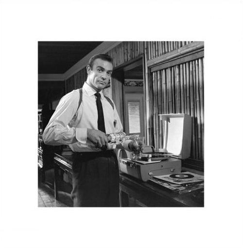 A man wearing a dress shirt and tie stands indoors, pouring a James Bond - Vodka Martini from a MEZ 245 bottle. He is next to an open record player with a vinyl record on it, in a room with wooden paneling. The setting feels vintage, yet accented by Pop Culture Themed Art Prints that lend it a unique flair.