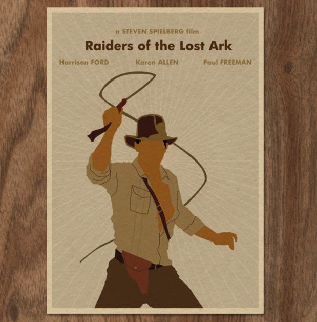 Poster for the film "Raiders of the Lost Ark Monster Gallery" featuring a stylized silhouette of a man in a fedora hat, holding a whip. The background is beige with the title and actors' names: Harrison Ford, Karen Allen, and Paul Freeman, along with "a Steven Spielberg film." The brand name Blue Dog Posters is also prominently displayed.
