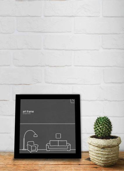 A minimalist **Classic Frame Square 21 x 21cm Black** from **Blue Dog Posters** showcasing a black and white sketch of a living room scene, featuring a couch, floor lamp, and small picture, placed on a wooden surface. To the right of the frame is a small potted cactus in a woven basket. The background is a white brick wall.